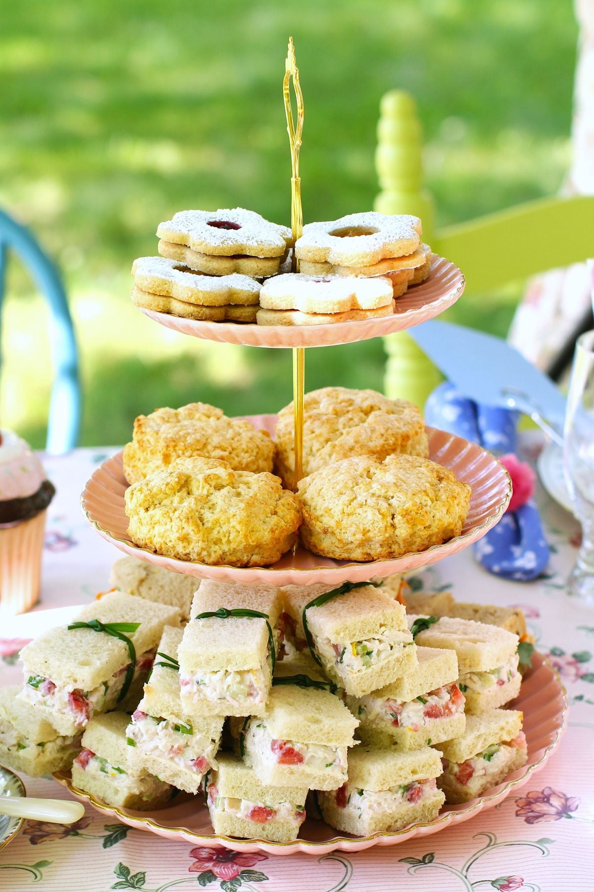 A 3-tiered cake stand with Strawberry Chicken Salad Tea Sandwiches on the bottom, scones in the middle, and cookies on the top.
