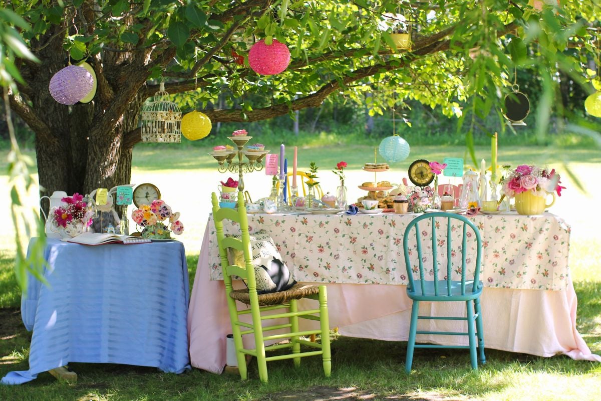 A large and a small tea table set with china, clocks, and whimsical flowers and treats, a green and blue chair, under the boughs of a pear tree hung with round coloured lanterns, clocks, and birdcages.