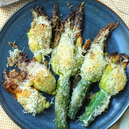 Square thumbnail photo of blue plate with 3 baked stuffed zucchini blossoms and 3 that are attached to small zucchinis, covered in Parmesan cheese.