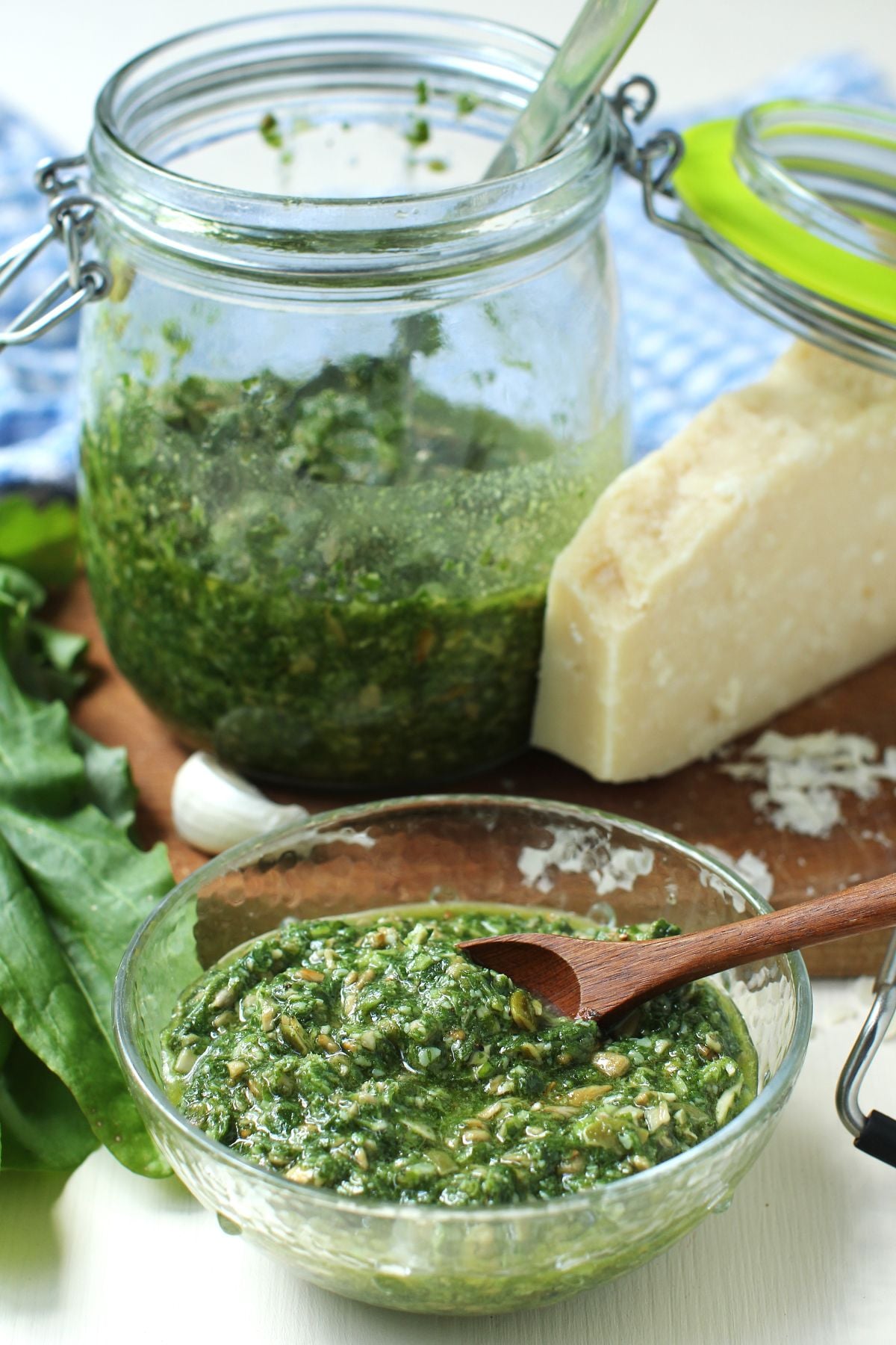 Small bowl of zucchini pesto in front, jar of it behind, sitting on a cutting board, with a wedge of Parmesan cheese beside it.