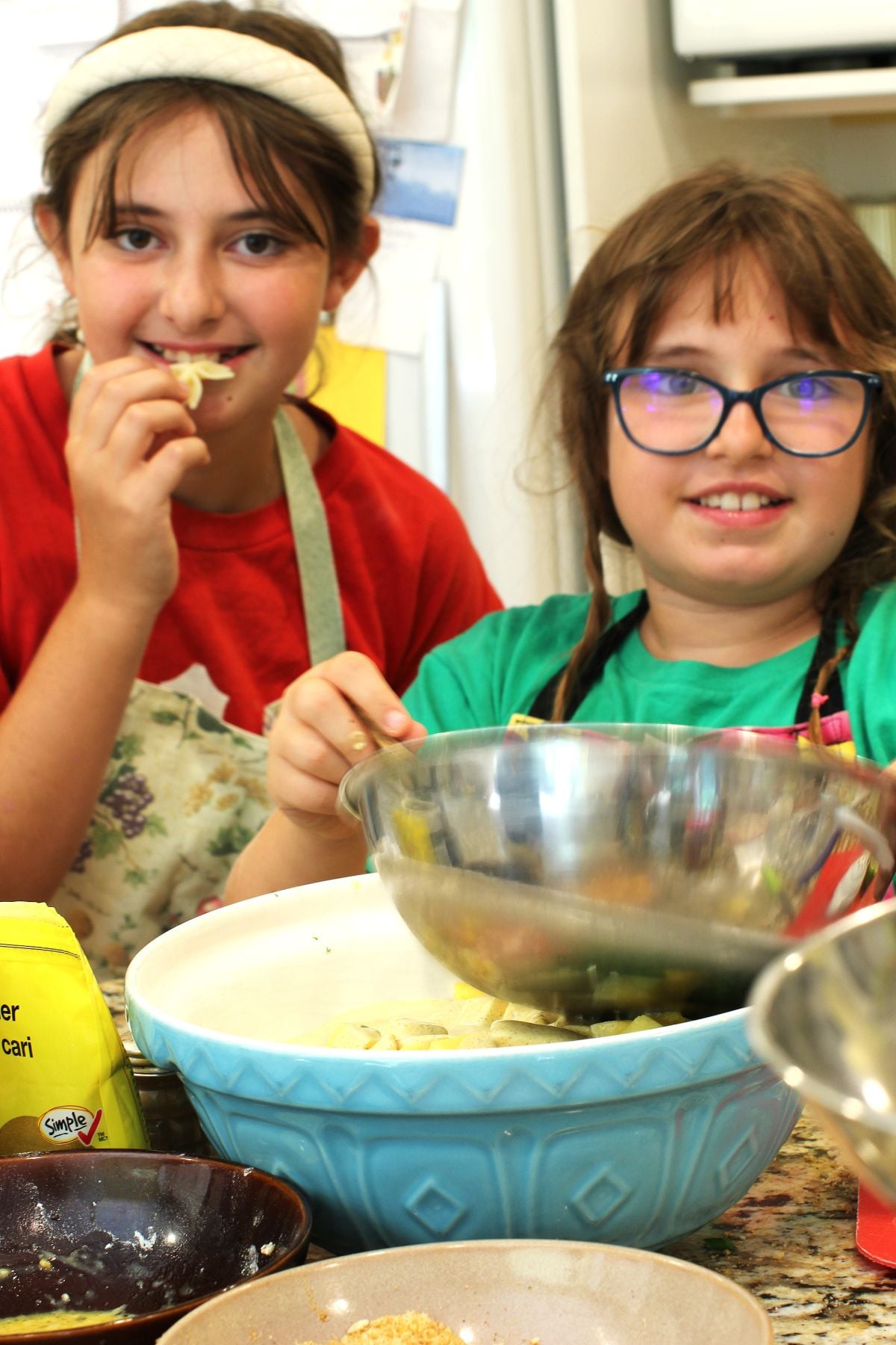 Willow and Ivy in the Kitchen, Willow eating a noodle, Ivy holding a stainless steel bowl.