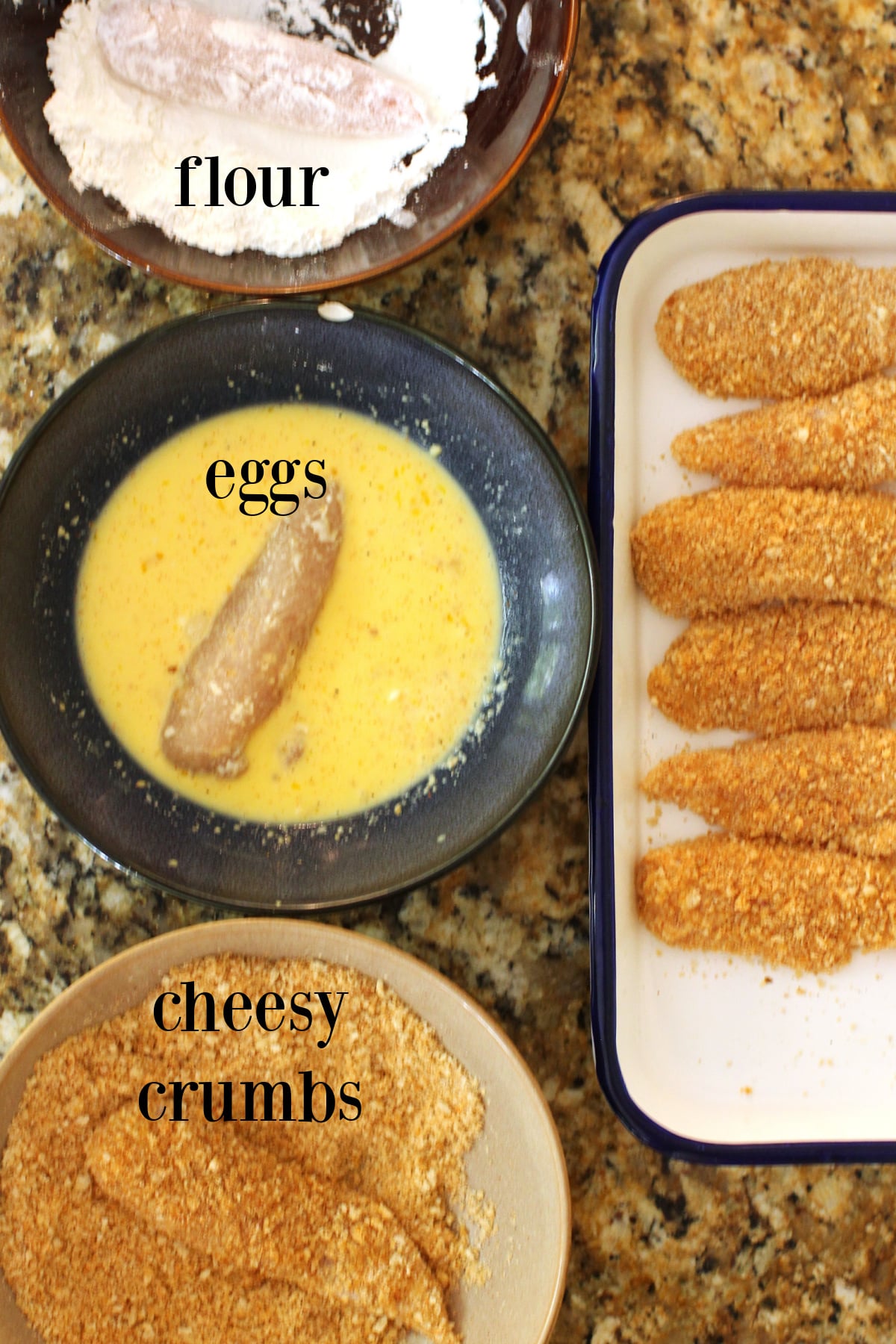Three bowls containing, flour, eggs, breadcrumbs, with chicken pieces in them, and a tray of the breaded fingers.