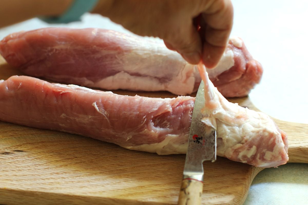 One hand lifting up the silverskin, while the knife slides along and cuts off the rest on a pork tenderloin.