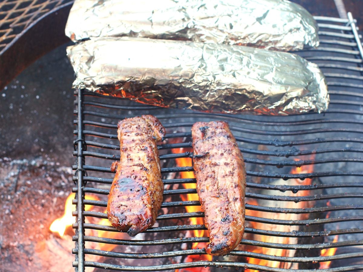 Two grilled tenderloins laying on a grill rack over the campfire, next to two foil packets of potatoes.