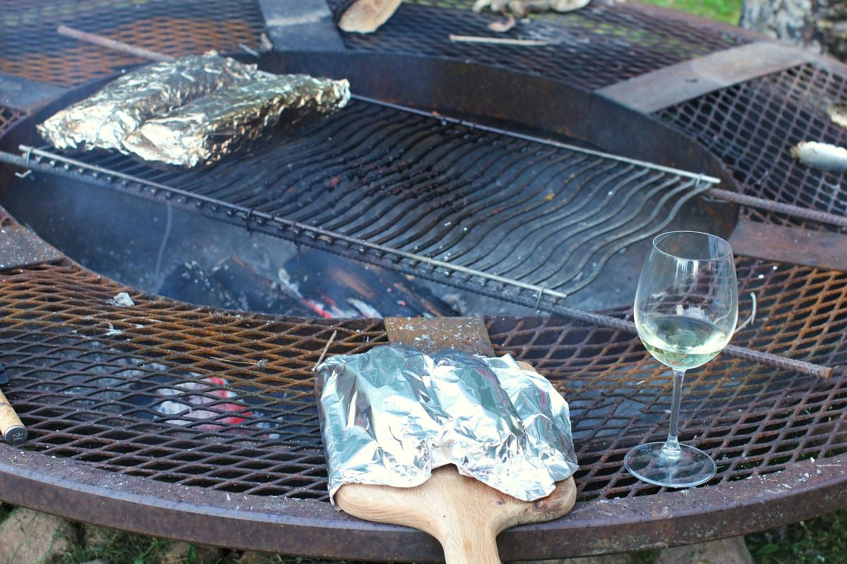 Tenderloins are laying on a cutting board, covered in foil, on the fire pit, with a glass of wine beside.