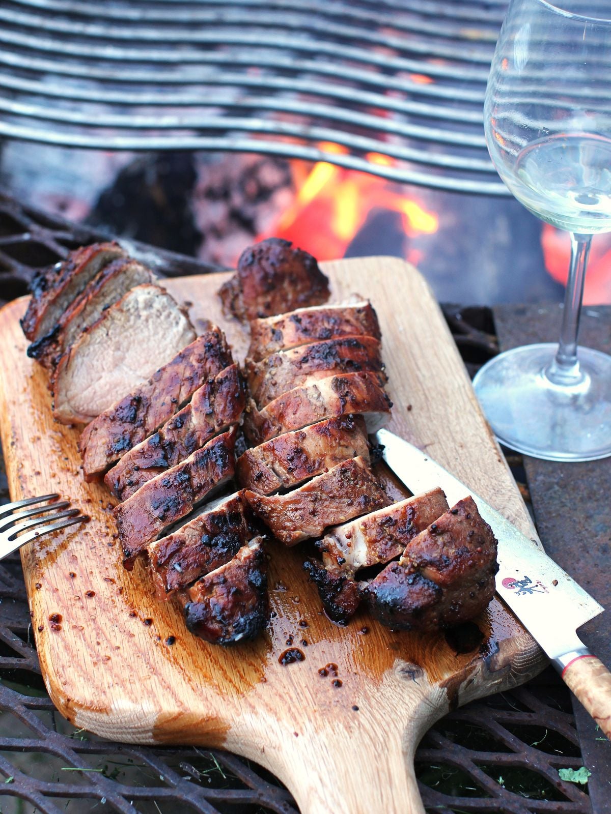 A wooden cutting board with two sliced, grilled tenderloin on them, the grill with some fire under it behind.